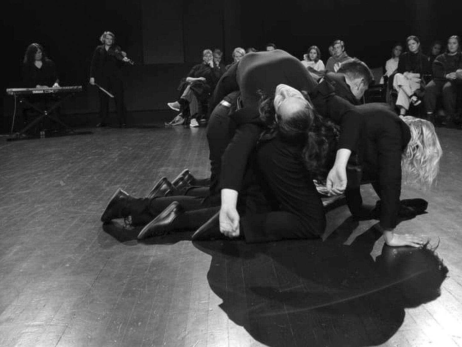 A black-and-white photograph shows an arrangement of dancers on the wooden floor of a performance space, with spectators in the background. Three dancers are positioned on their hands and knees, with a fourth dancer draped over them.