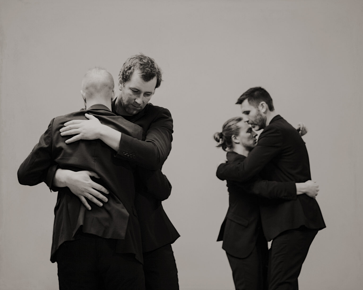 In a black-and-white photograph, two pairs of men wearing dark suits embrace, dancing a tango.
