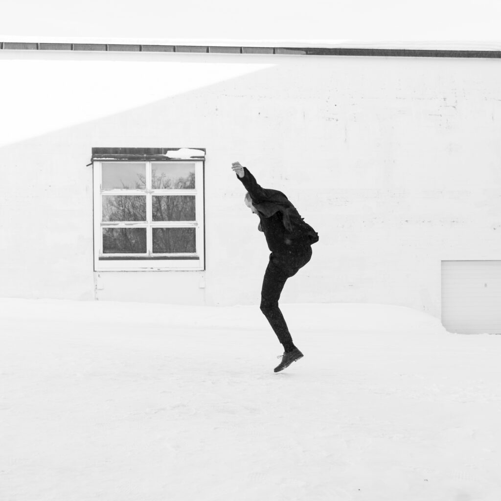 A black-and-white photograph shows a dancer in a black suit leaping in a snowy courtyard.