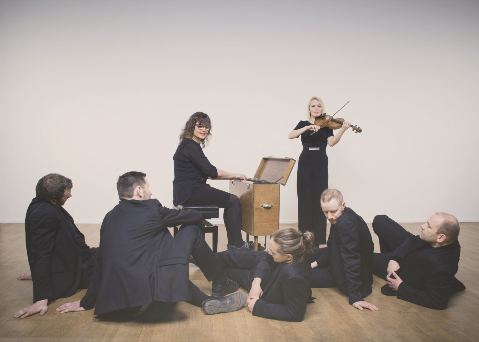 Five dancers in black suits lounge on the floor in front of two musicians—one plays a keyboard in a wooden box, while the other plays a violin.
