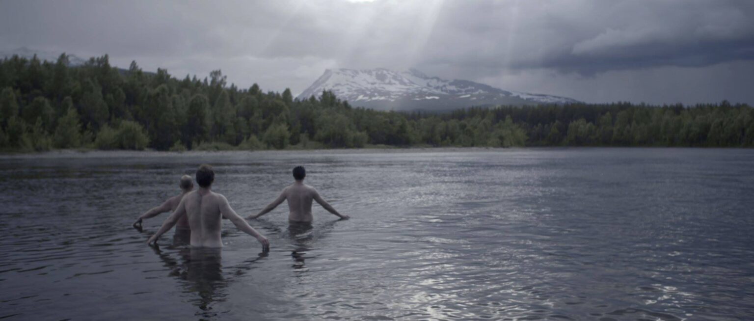 Three men wade into a lake, looking out onto forests and a mountain in the distance. Sunbeams cut through a cloudy sky.